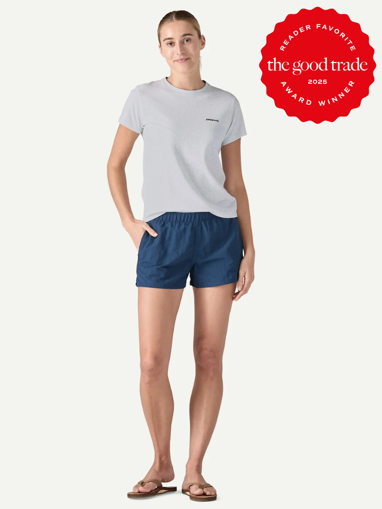 A woman stands wearing a light gray t-shirt, navy shorts, and sandals. The image features a red "The Good Trade 2025 Award Winner" badge in the top right corner.