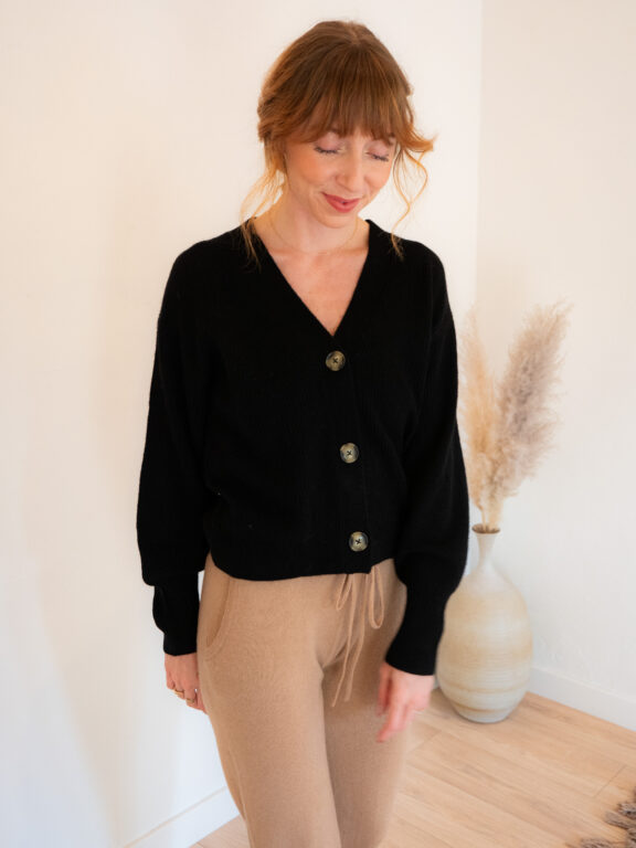 Woman with light brown hair wearing a black button-up cardigan and beige drawstring pants stands indoors near a vase with pampas grass.