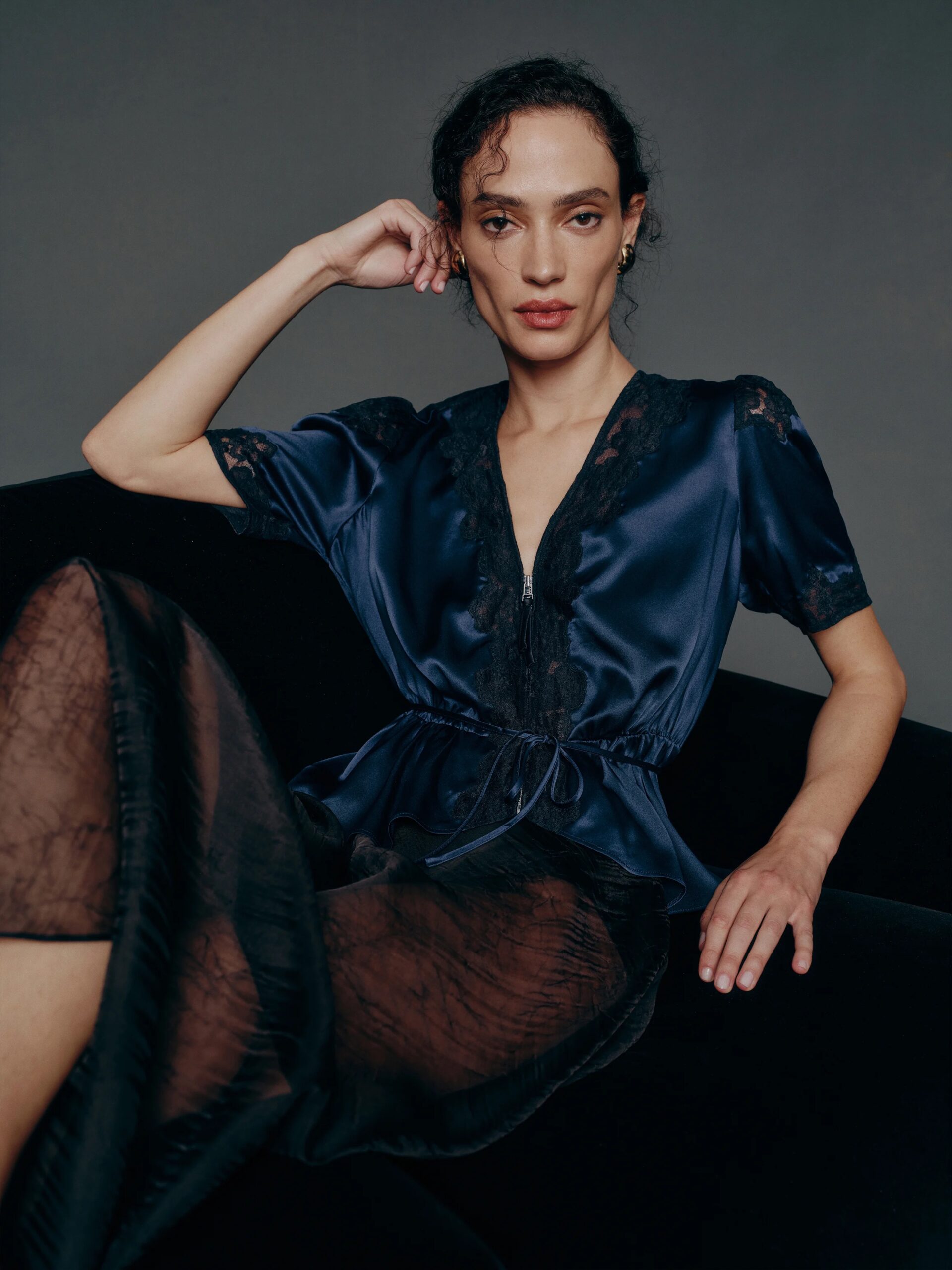 A person with curly hair sits on a black chair, wearing a navy satin blouse with lace details and a sheer black skirt, posing against a dark background.