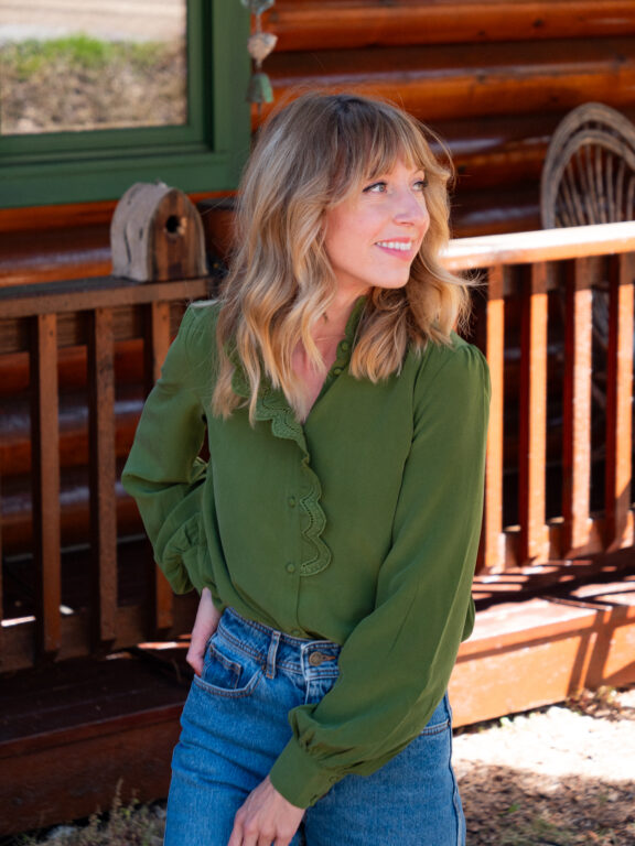 A woman with blonde hair wearing a green blouse and blue jeans stands outside in front of a wooden railing, looking to her left.