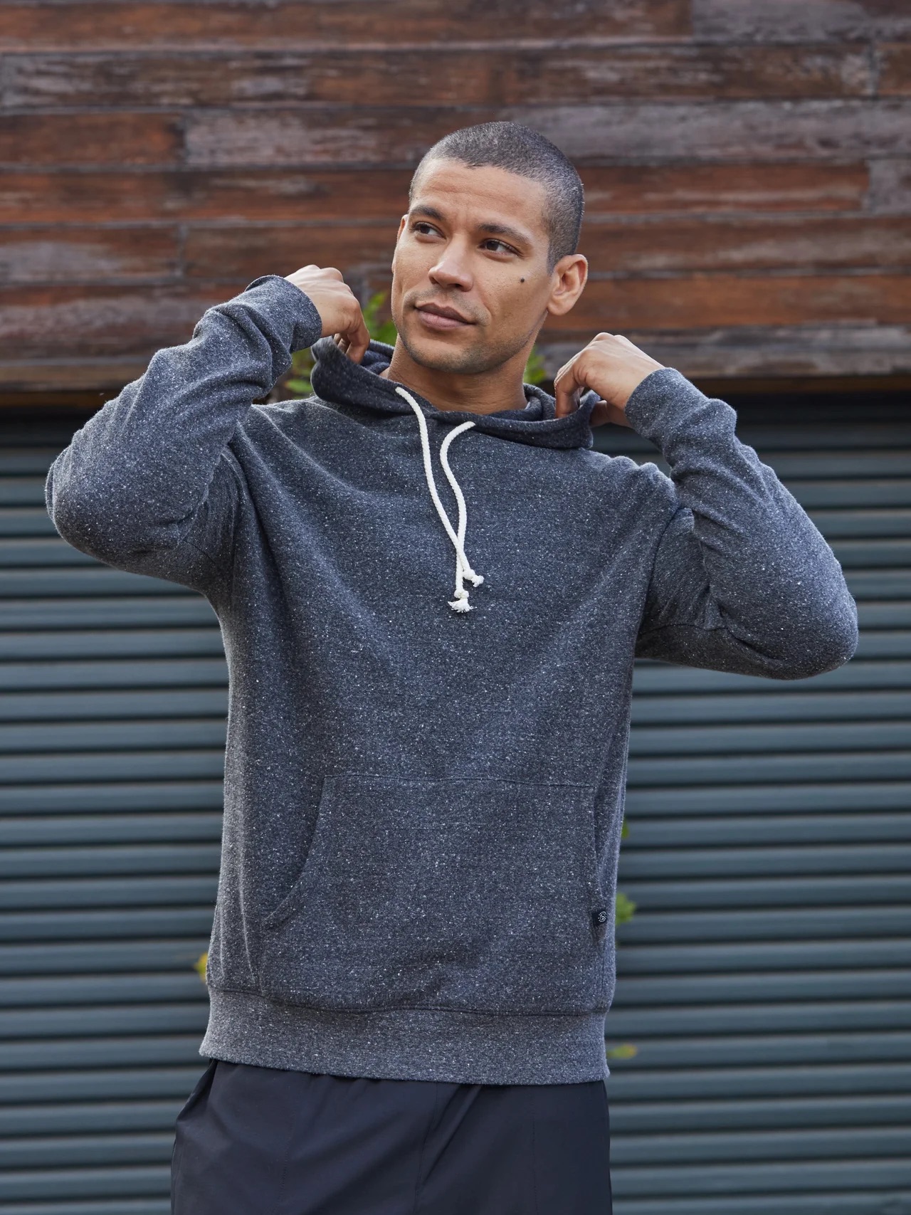 A man stands outdoors in front of a wooden wall and closed garage door, adjusting the hood of his dark gray hoodie.