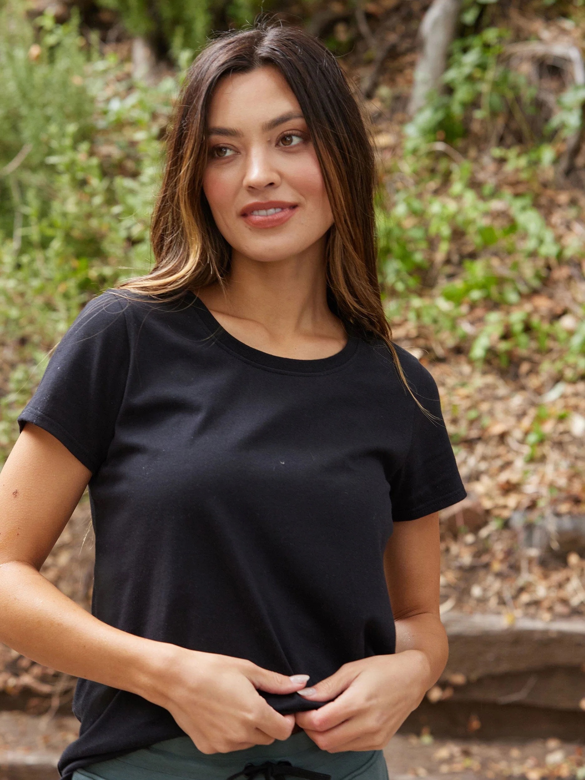 A woman with long brown hair stands outdoors wearing a black t-shirt, looking slightly to the side, with a background of greenery and fallen leaves.