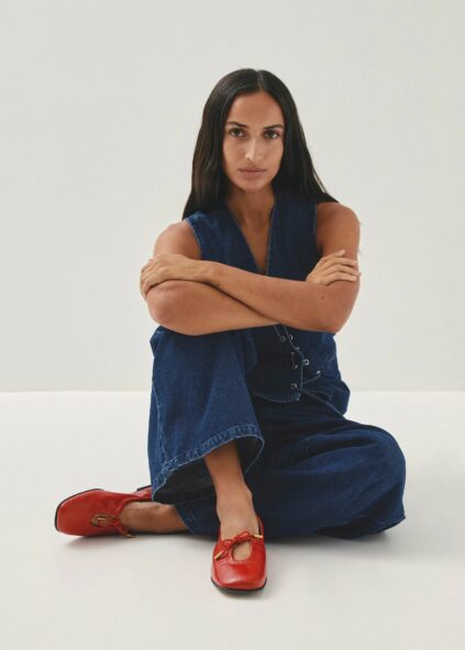 Person with long dark hair sits cross-legged on the floor, wearing a blue sleeveless jumpsuit and red shoes, against a plain white background.