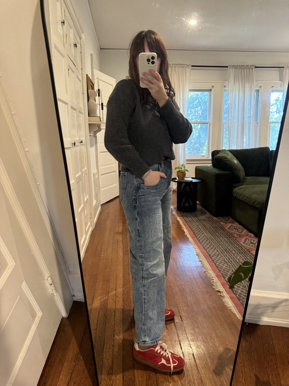 Person taking a mirror selfie in a room with wooden floors, wearing a dark sweater, blue jeans, and red sneakers. The room has white walls, windows with curtains, and a green sofa.