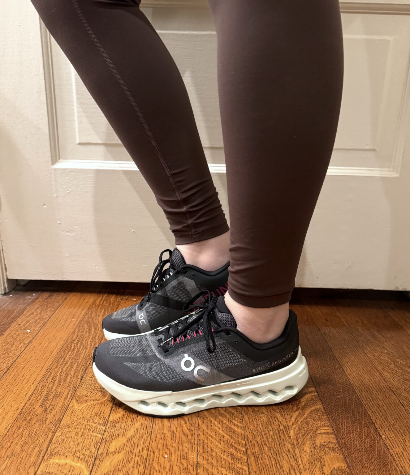 Person wearing black running shoes with white soles and brown leggings stands on wooden floor near a white door.
