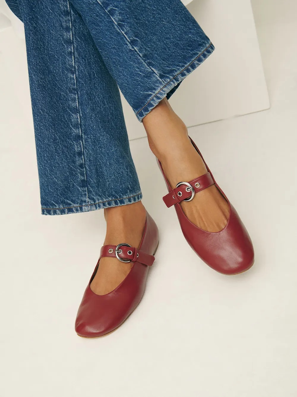 Person wearing red leather Mary Jane flats with buckle straps, paired with blue denim jeans.