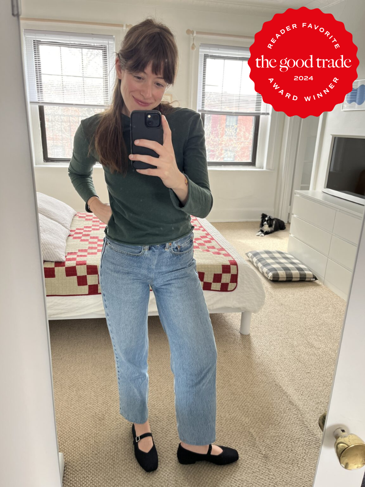 Woman taking a mirror selfie in a bedroom, wearing a green sweater and jeans. There's a bed with a red-and-white quilt and a cat on a rug in the background. "The Good Trade Award Winner" badge.