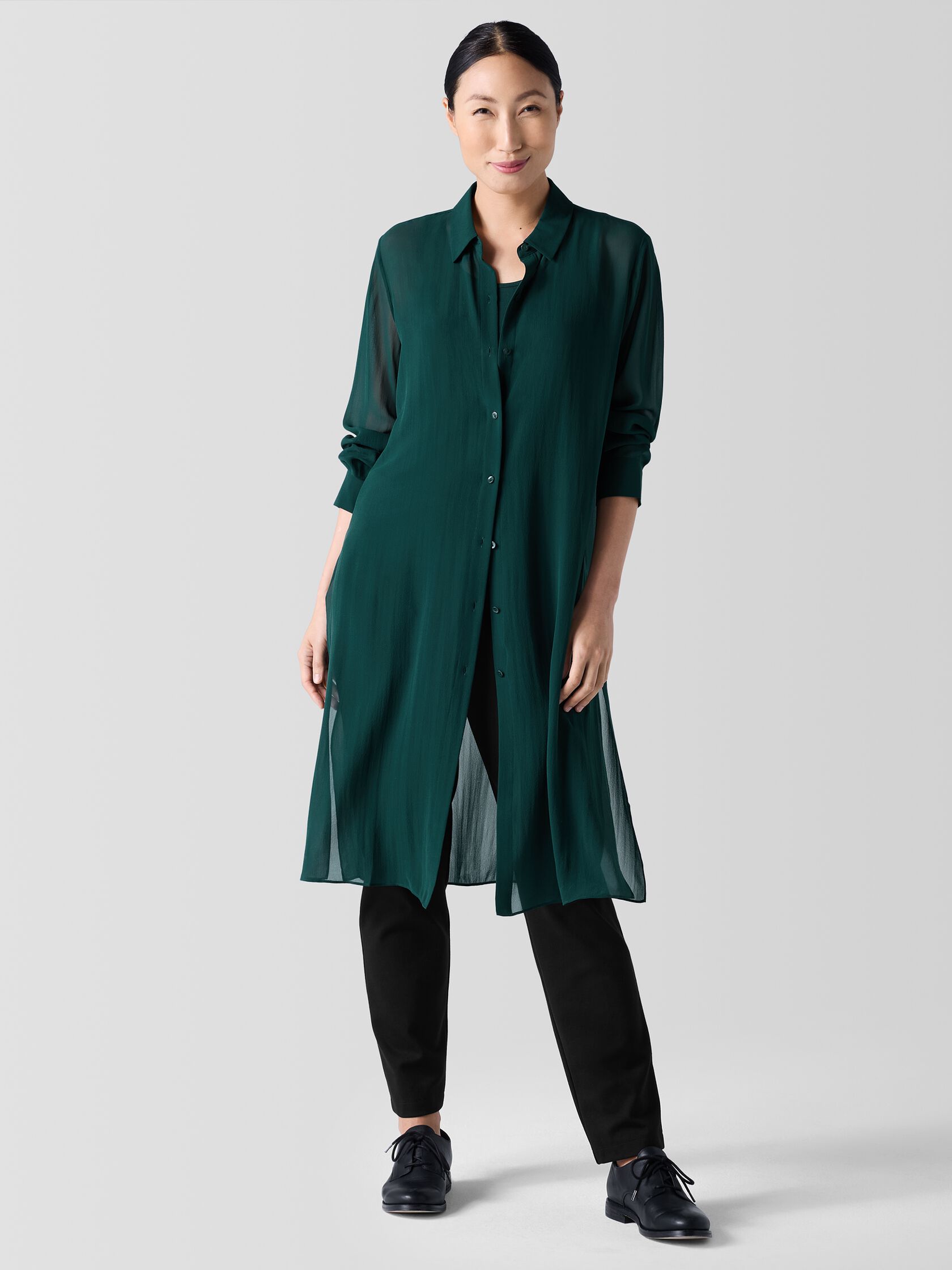 A person stands against a light background, wearing a dark green long button-up tunic over black pants and black shoes.