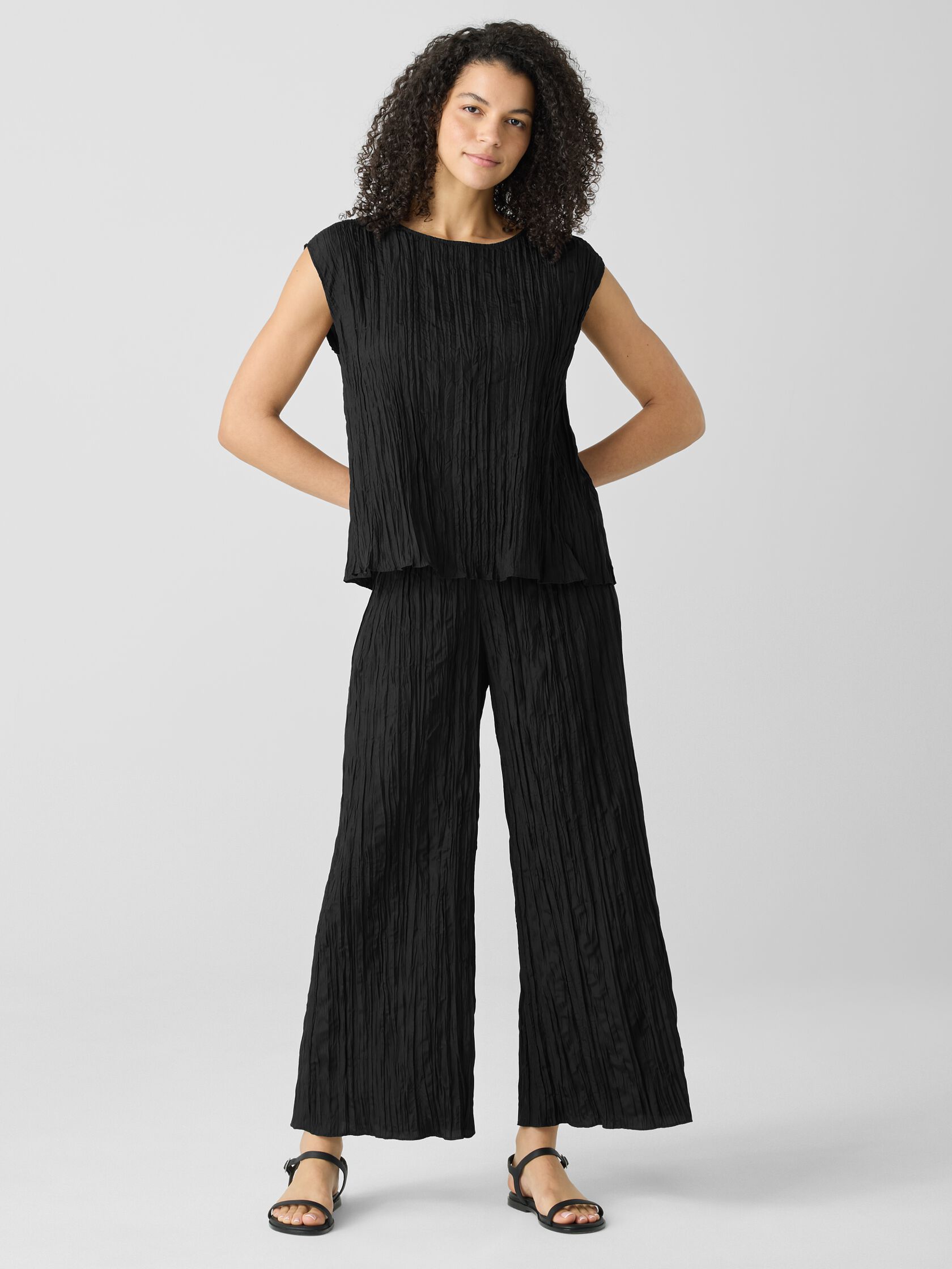 A woman stands against a plain background wearing a black, loose-fitting, crinkled two-piece outfit with wide-leg pants and a sleeveless top, paired with black sandals.