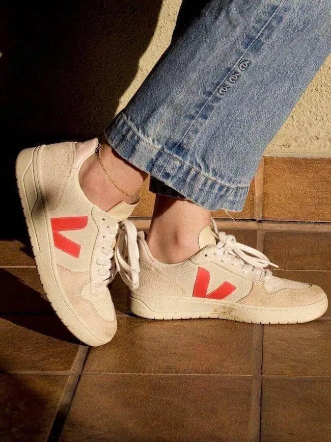 Person wearing beige sneakers with red accents and blue jeans standing on a tiled floor.