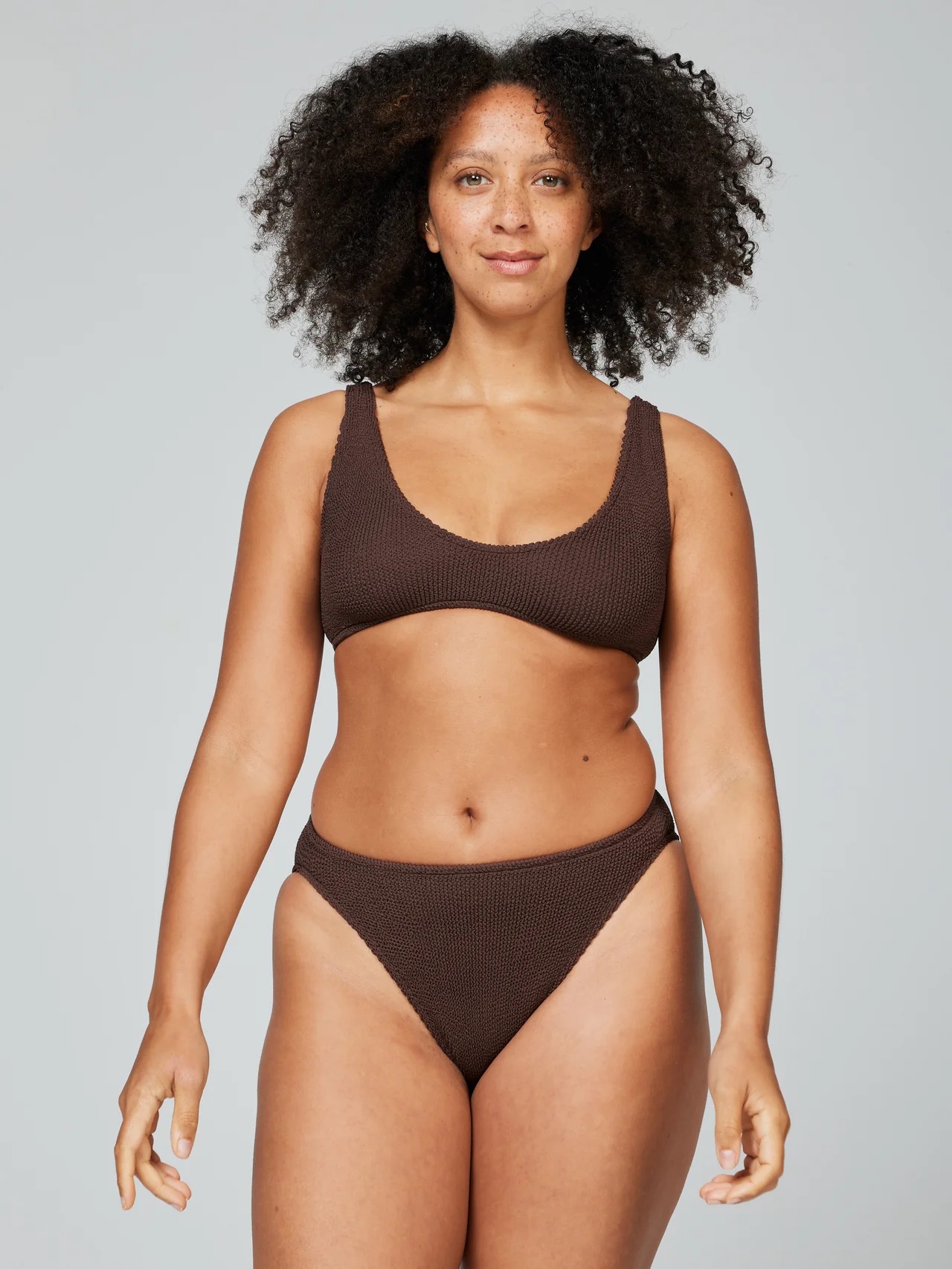 A person with curly hair stands confidently in a brown textured bikini.