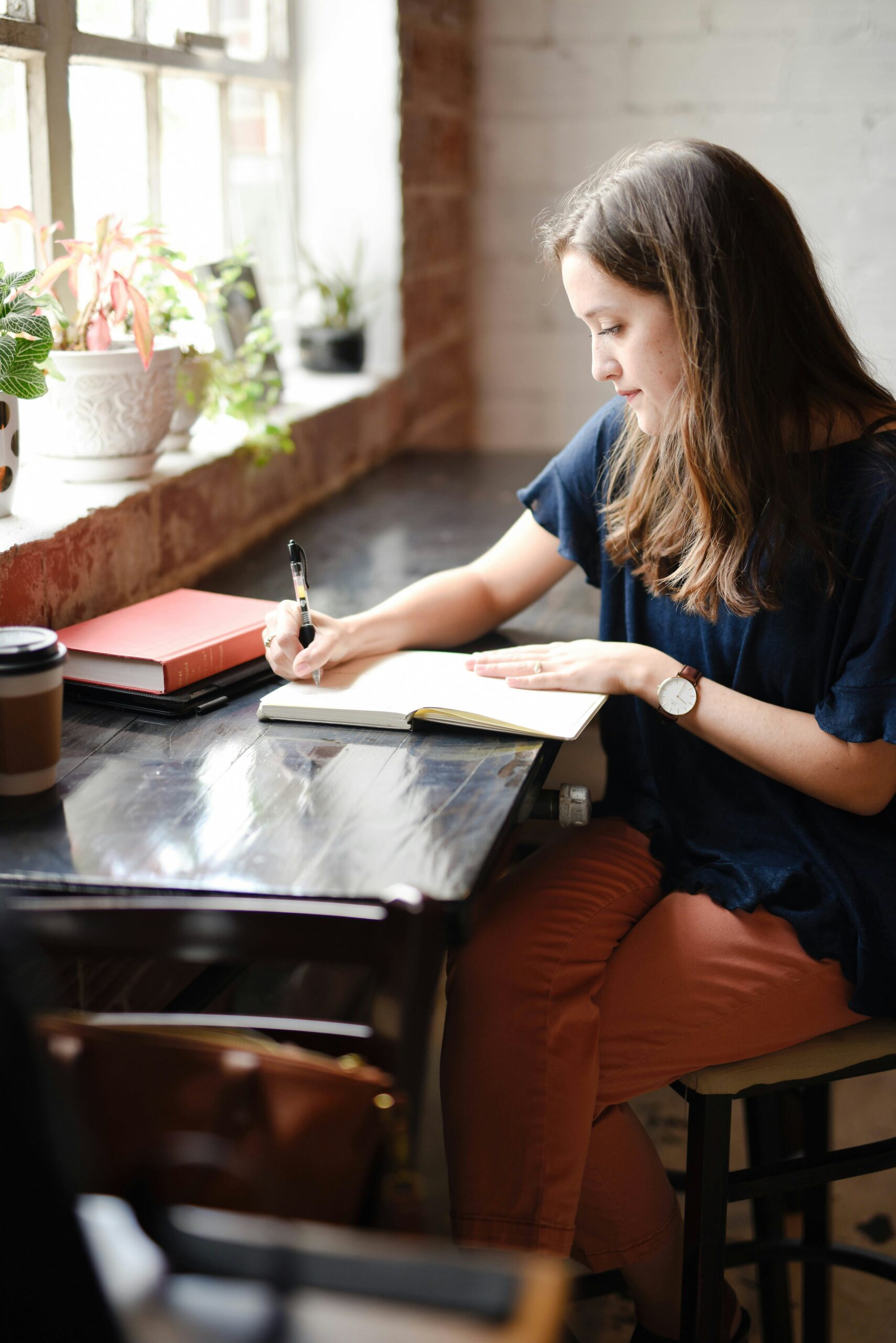 A woman writes in a notebook at a cafe table with a coffee cup, a red book, and potted plants on the windowsill nearby.