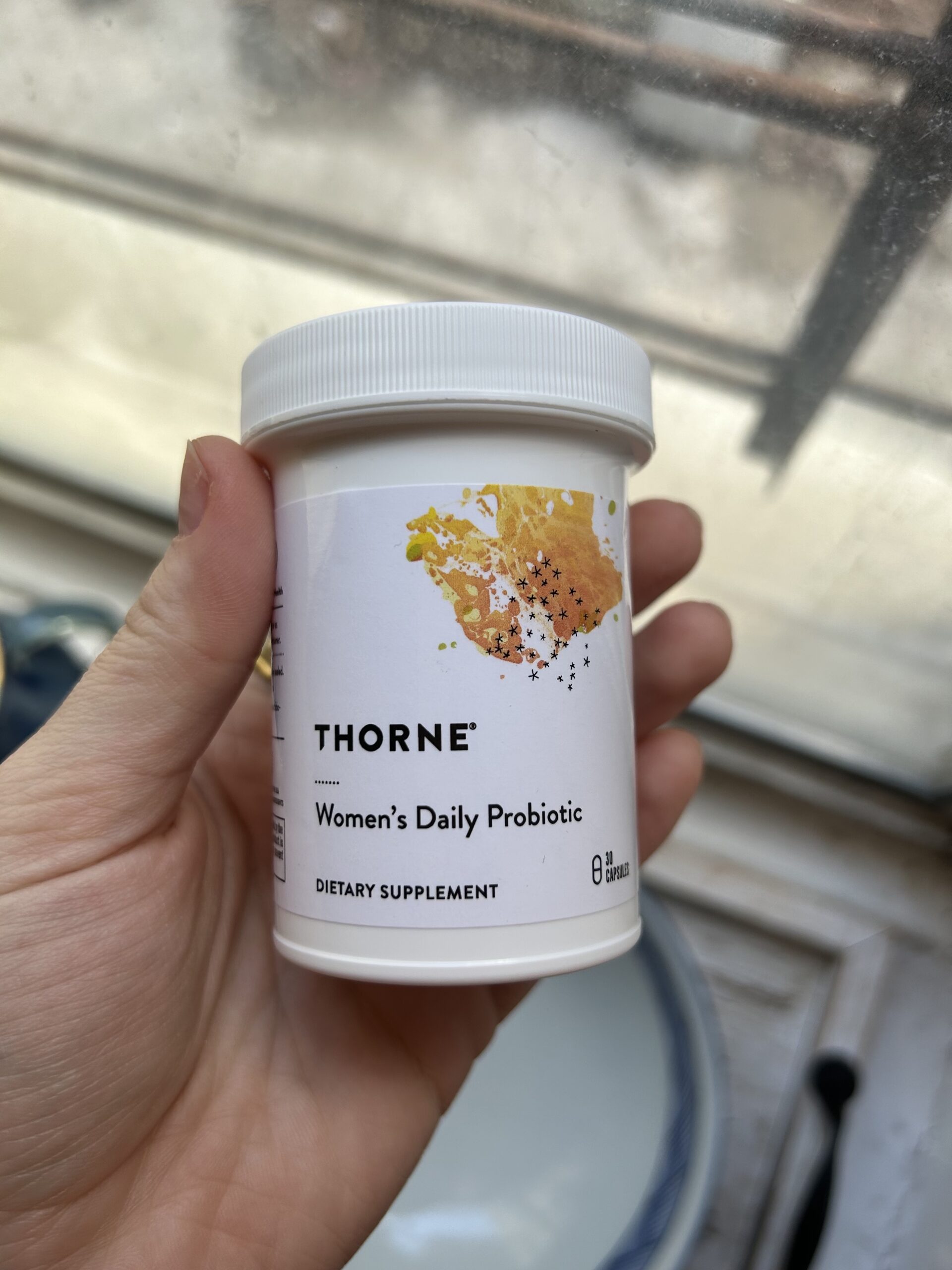 A hand holds a white container of Thorne Women's Daily Probiotic dietary supplement in front of a window.