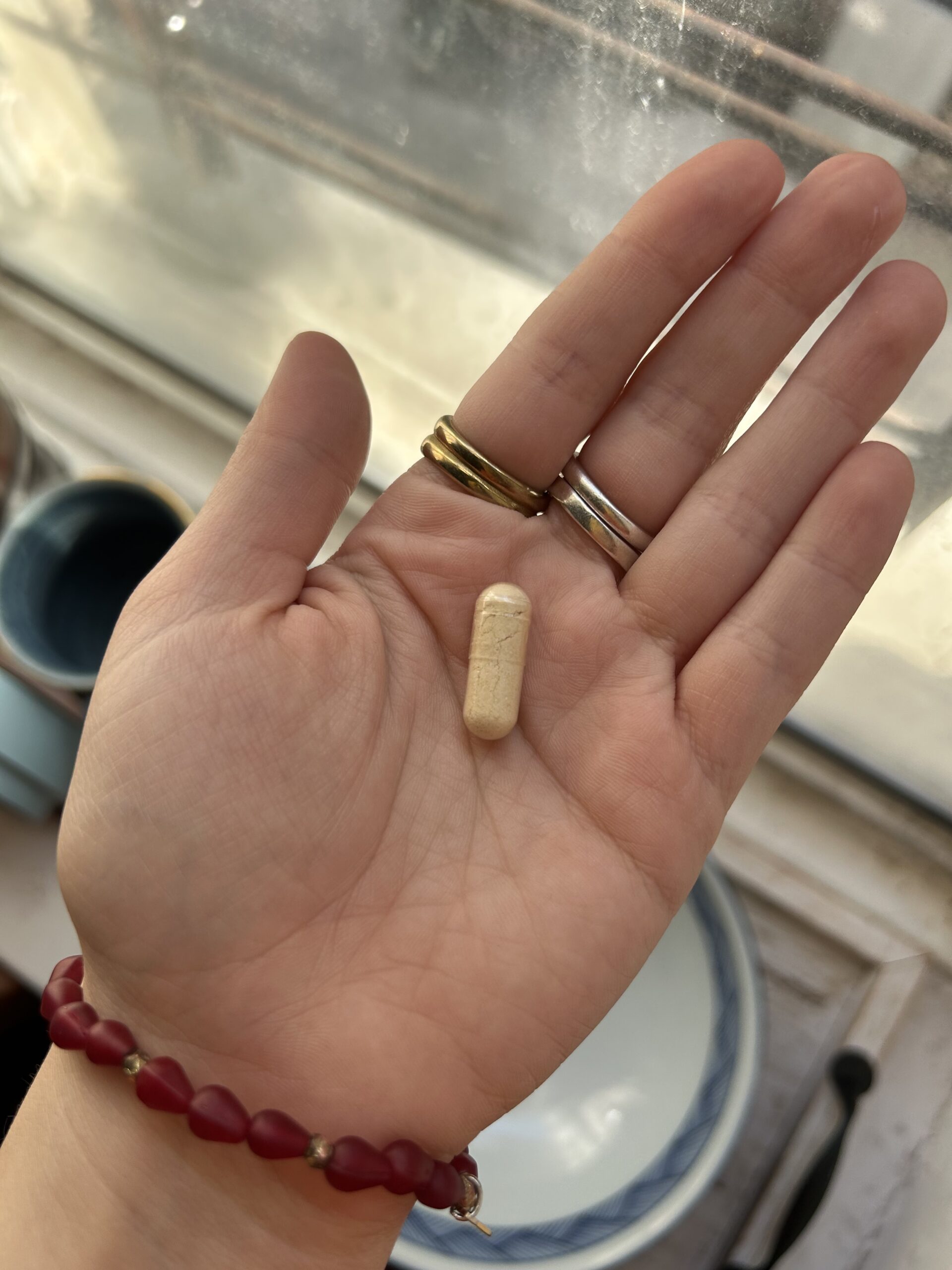 A hand with three rings and a red beaded bracelet holds a single beige capsule pill above a few bowls and cups.