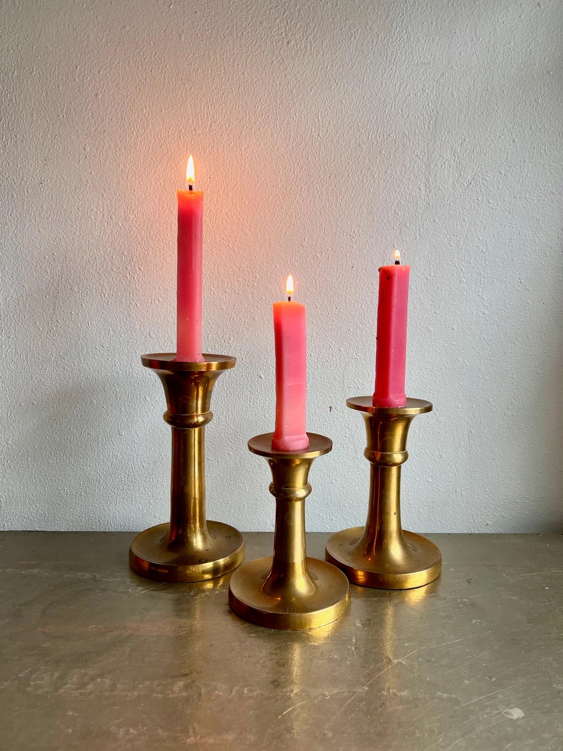 Three lit pink candles of varying heights stand on a surface in brass holders against a neutral background.