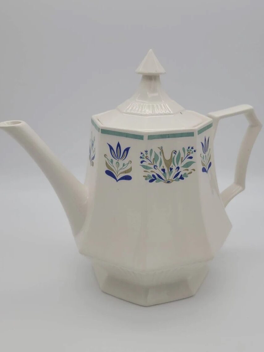 A white ceramic teapot with a hexagonal shape, featuring blue and green floral designs. It has a pointed lid and a distinct handle and spout.