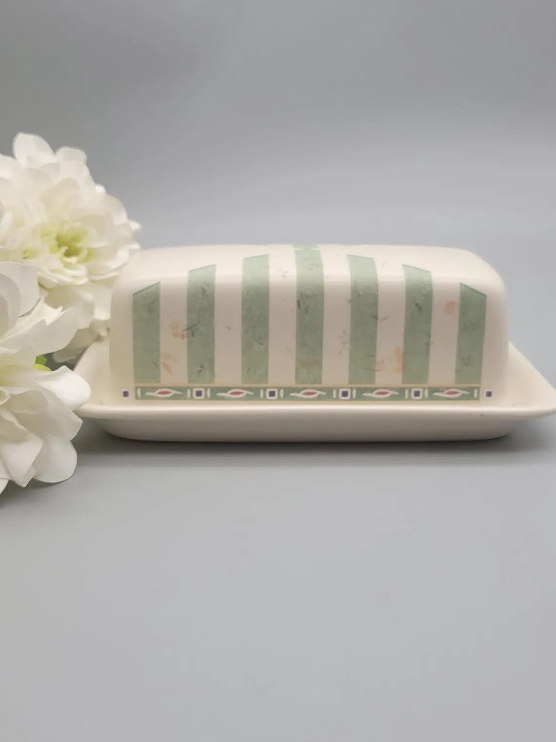 A butter dish with a green stripe and pattern design sits on a light grey surface, accompanied by white flowers on the left.
