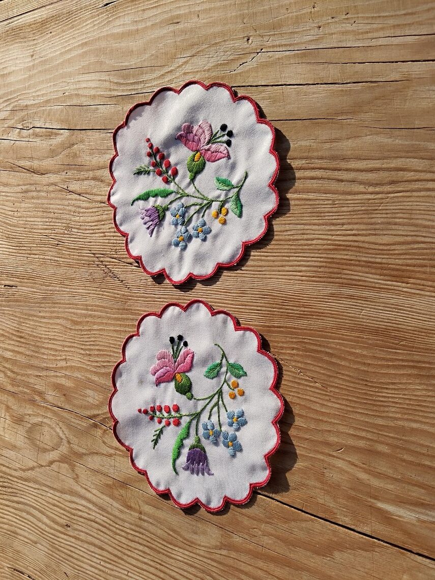 Two round embroidered doilies with floral patterns, featuring pink, purple, and red flowers, are placed on a wooden surface.