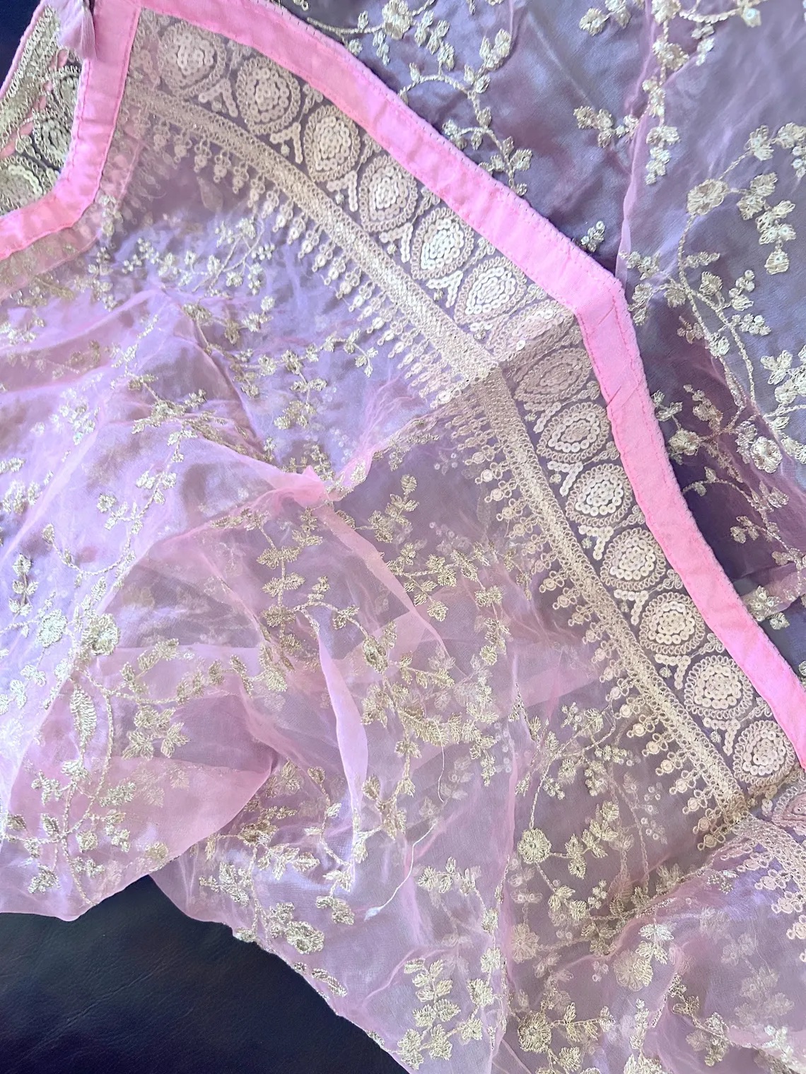 A close-up of a sheer pink fabric with intricate floral embroidery and a decorative border featuring silver patterns and a pink edge.