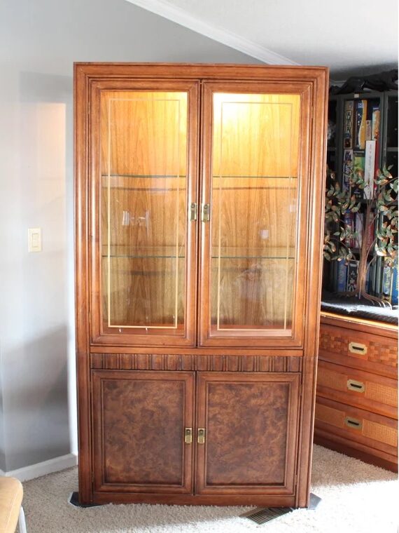 Wooden display cabinet with glass doors and shelves, standing in a room with a light gray wall and a chest of drawers nearby.