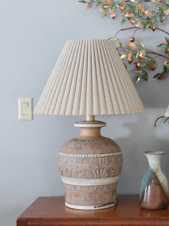 Two table lamps with pleated shades sit beside a small decorative vase on a wooden surface, with a metallic leaf wall decoration in the background.