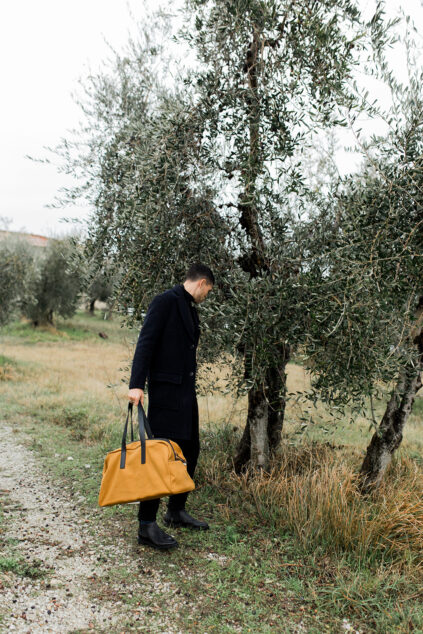 A person in a dark coat stands beside olive trees, holding a yellow bag, in an outdoor setting with grass and overcast skies.