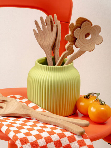 Wooden kitchen utensils in a green vase on an orange chair, with more utensils on an orange checkered cloth. Two orange tomatoes are next to the cloth.