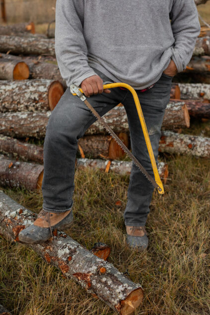 Person in grey sweater and jeans holds a yellow saw while resting a foot on a cut log; more logs are stacked in the background.