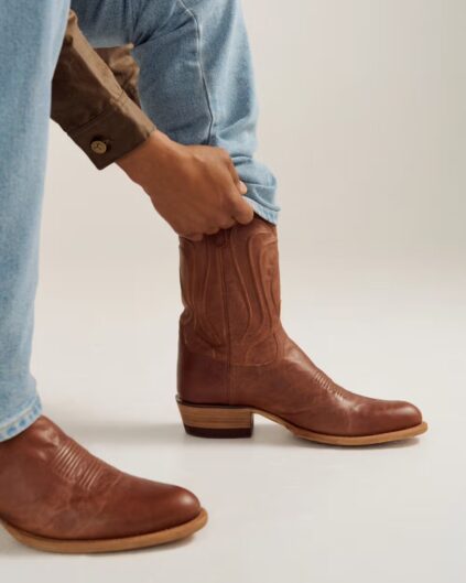 A person in light blue jeans and a brown shirt pulls on a brown leather cowboy boot, with another boot already on the other foot, against a plain background.