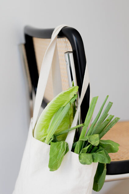 A canvas bag hangs on a chair, containing fresh green vegetables, including lettuce and scallions, against a neutral background.
