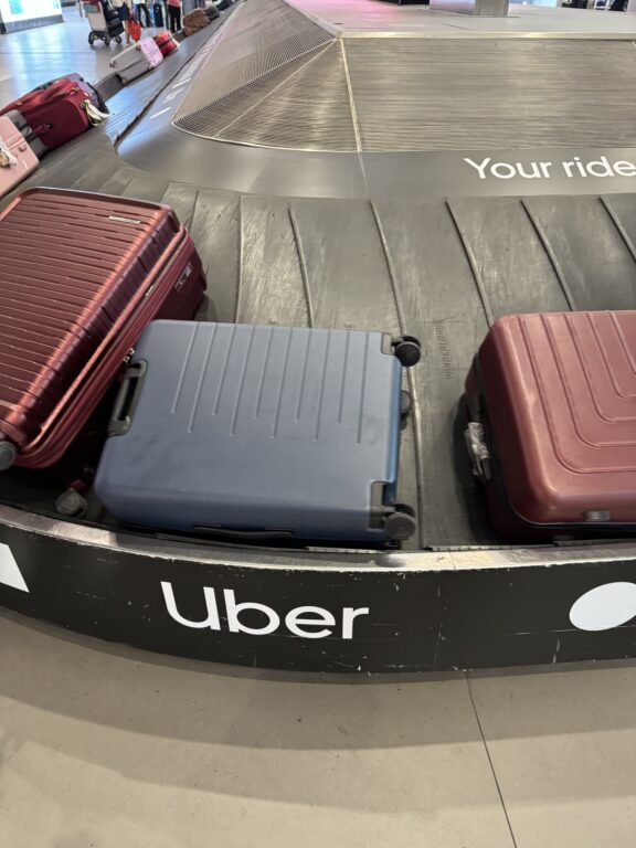 Several suitcases, including a blue one, are on an airport baggage carousel. The words "Uber" and "Your ride" appear on the carousel.