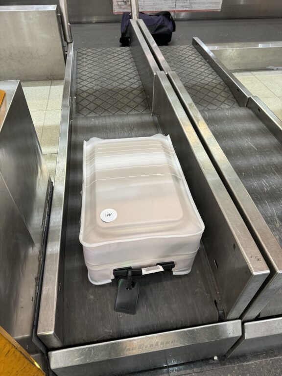 A white hard-shell suitcase with a luggage tag and a black item attached is placed on an airport baggage conveyor belt.