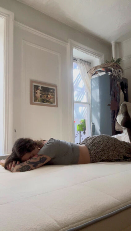 A person lies face down on a bed in a sunlit room with white walls, a window, a wardrobe, and a framed picture on the wall.