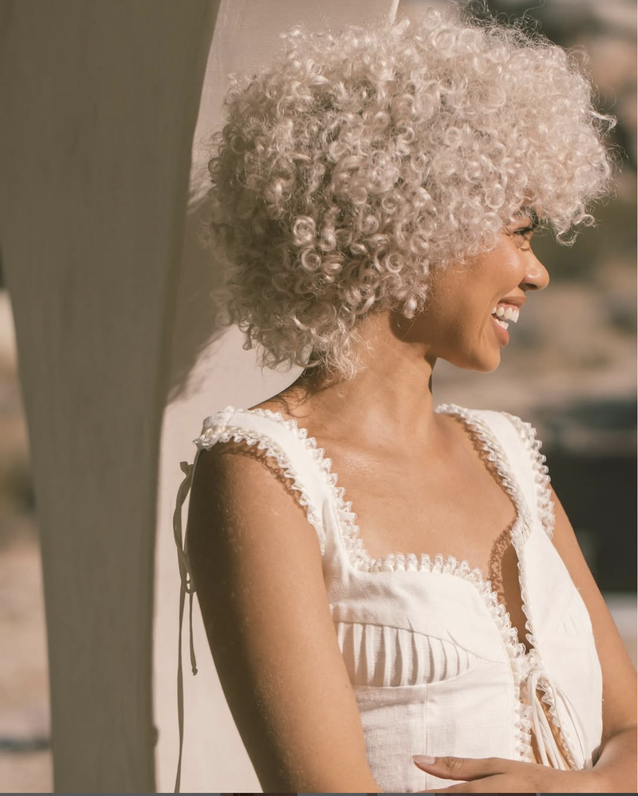 Person with curly, light hair smiles while standing outdoors in sunlight, wearing a sleeveless white top.