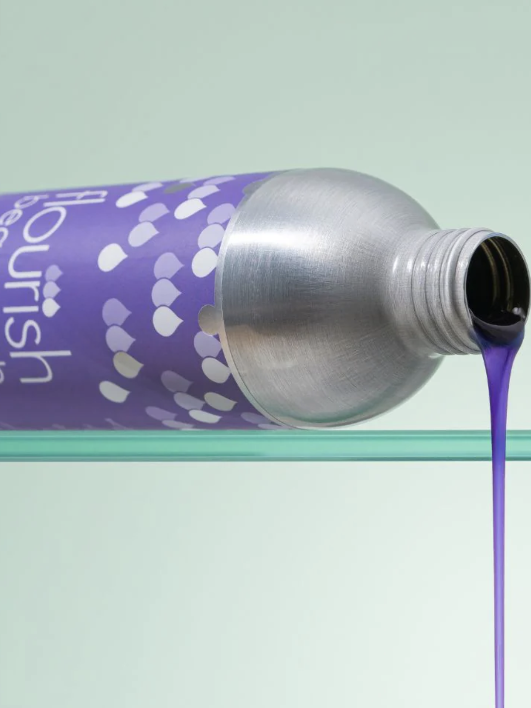 A purple liquid is pouring from a tilted aluminum tube with a purple label covered in small heart shapes, onto a glass surface.