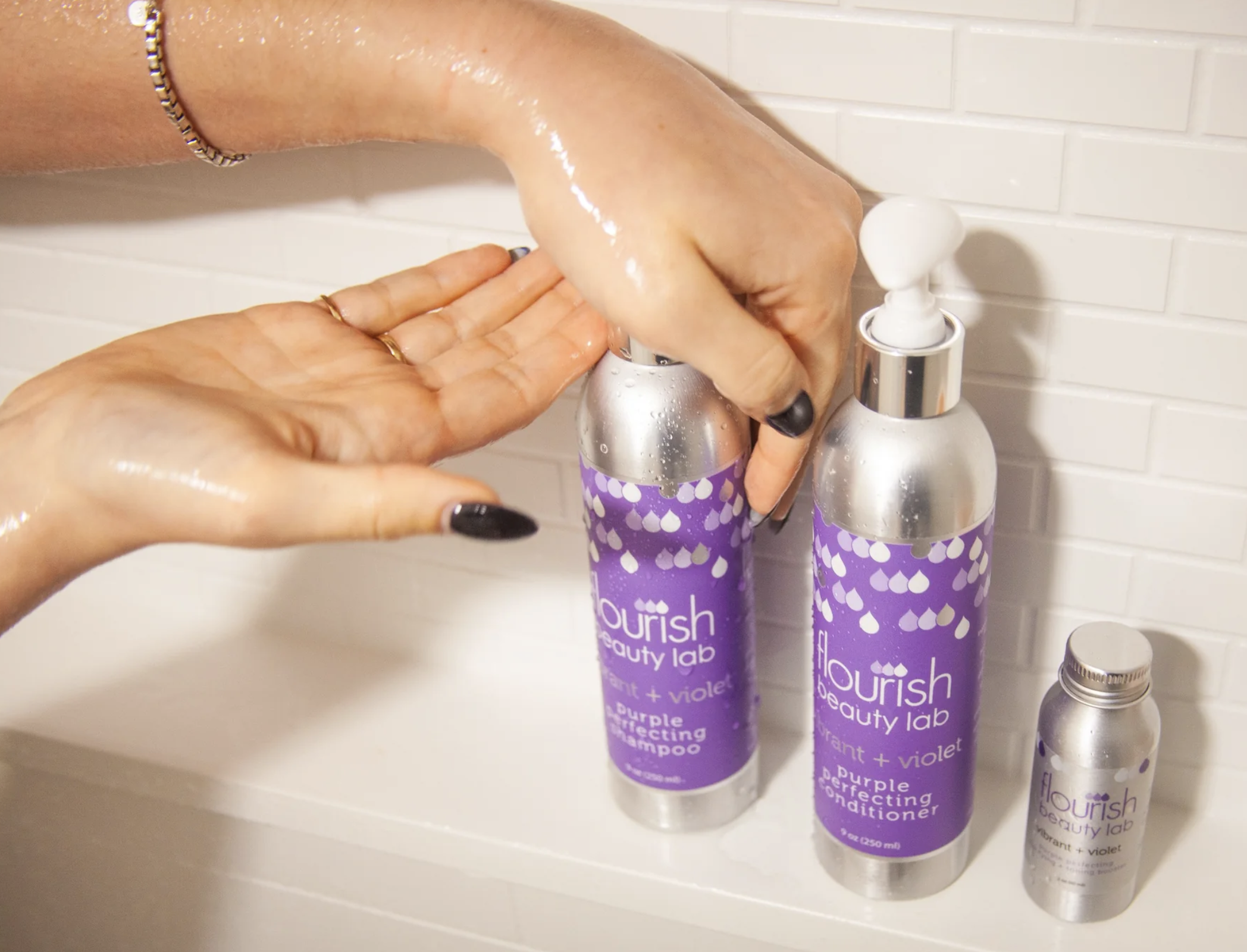 A person with wet hands dispenses shampoo from a silver bottle labeled "Nourish Beauty Lab," next to a matching conditioner bottle and a smaller silver container, in front of a tiled wall.