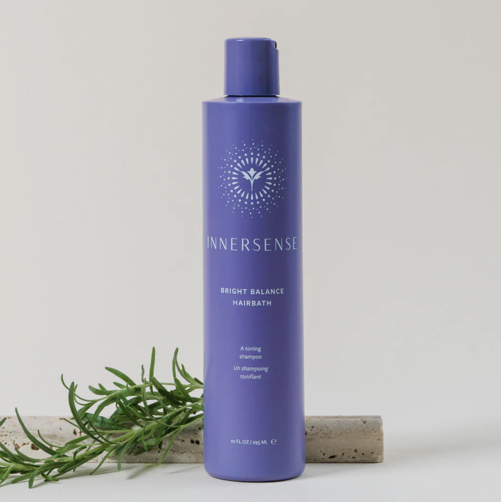 Purple bottle of Innersense Bright Balance Hairbath shampoo placed upright on a light surface with a sprig of rosemary nearby.