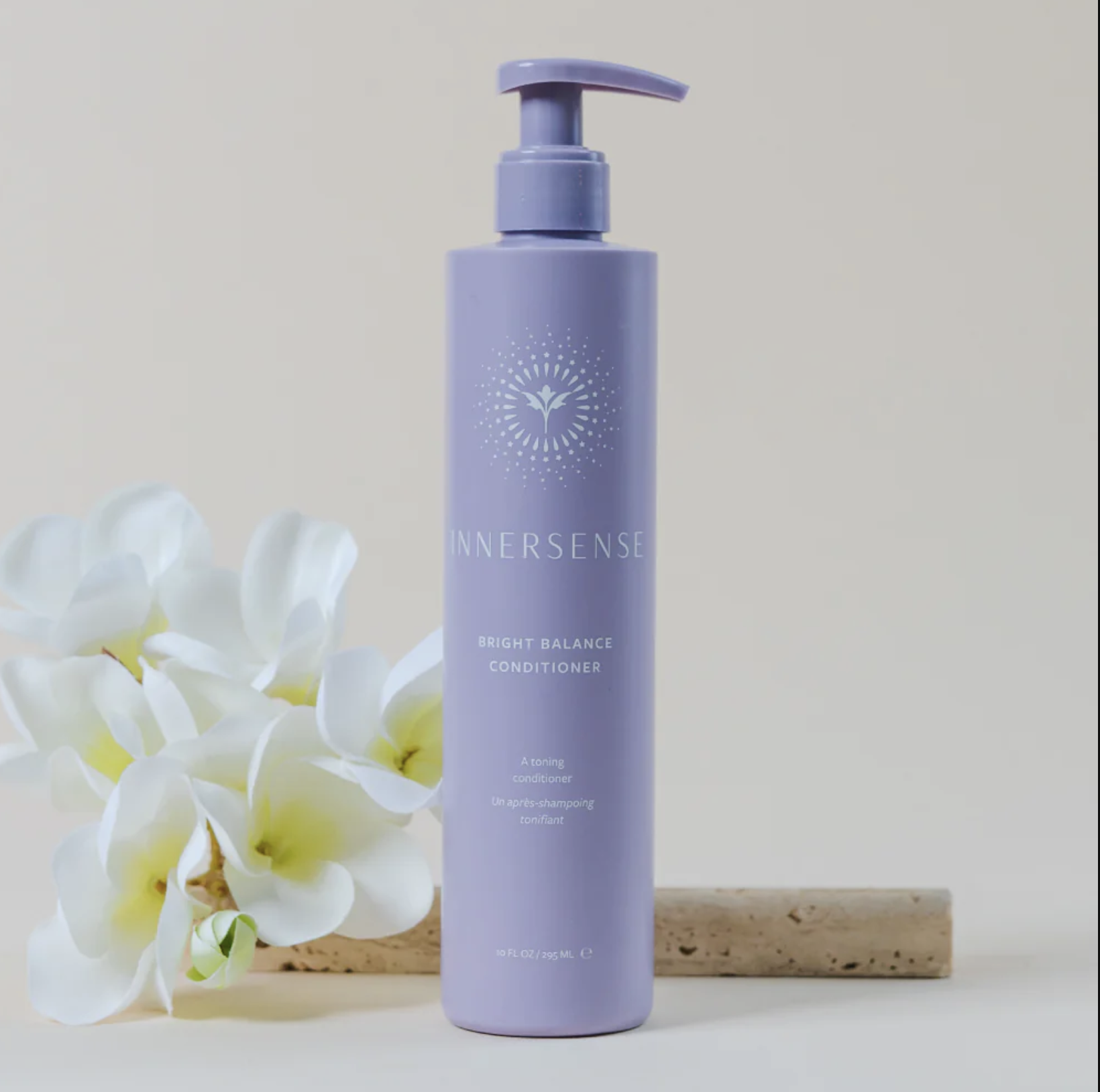 Purple bottle of Innersense Bright Balance Conditioner with pump nozzle, standing next to white flowers on a wooden surface.