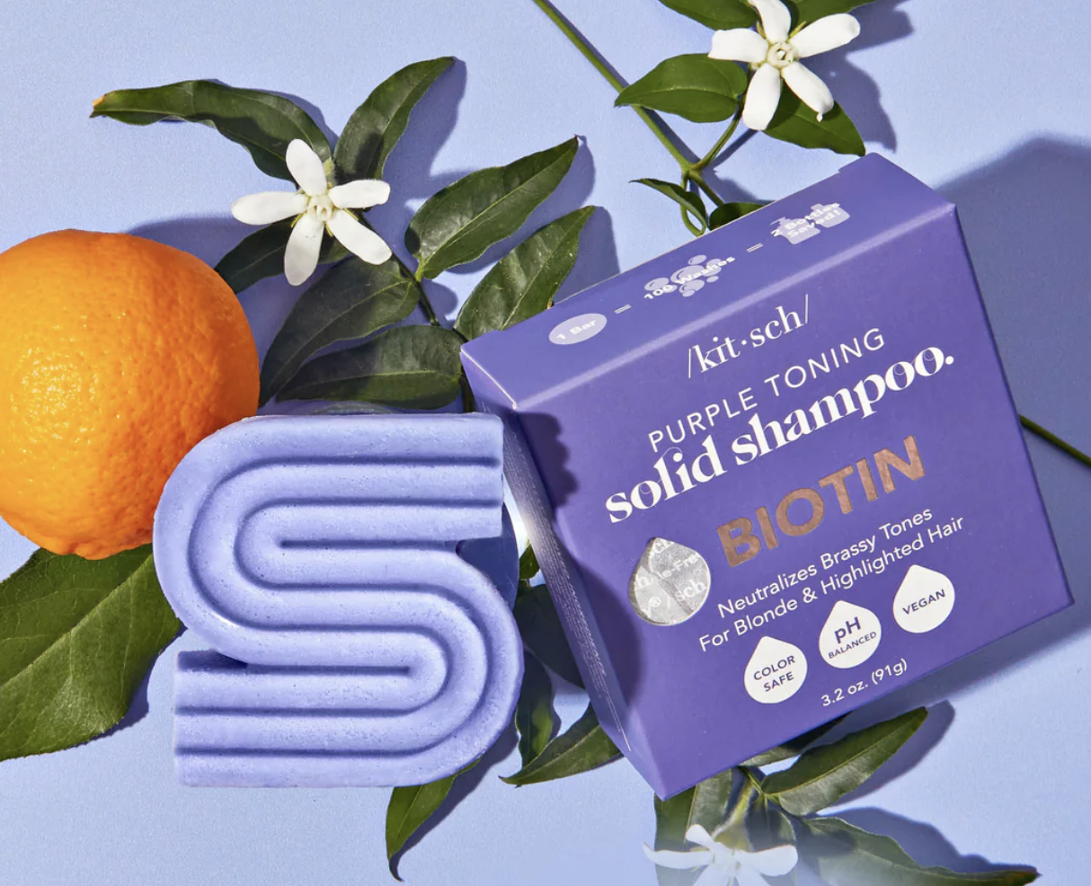 Purple toning solid shampoo and its box on a blue surface with an orange and green leaves. The shampoo is designed for blonde and highlighted hair, featuring biotin and vegan ingredients.