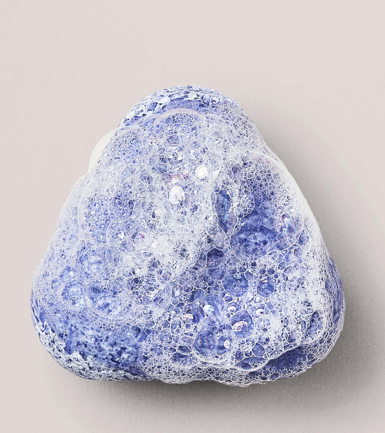 A lavender-colored soap bar covered in white, frothy bubbles against a light beige background.