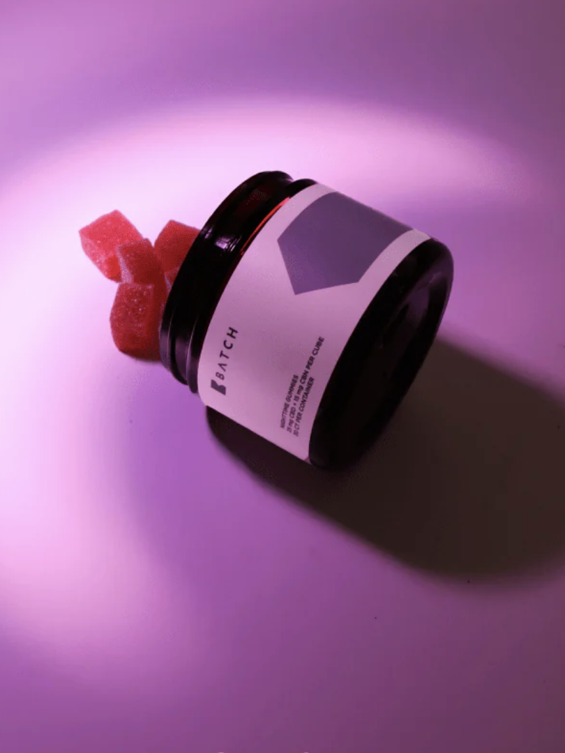 A dark jar labeled "Batch" lies on its side on a purple-lit surface, with several red gummy cubes spilling out.