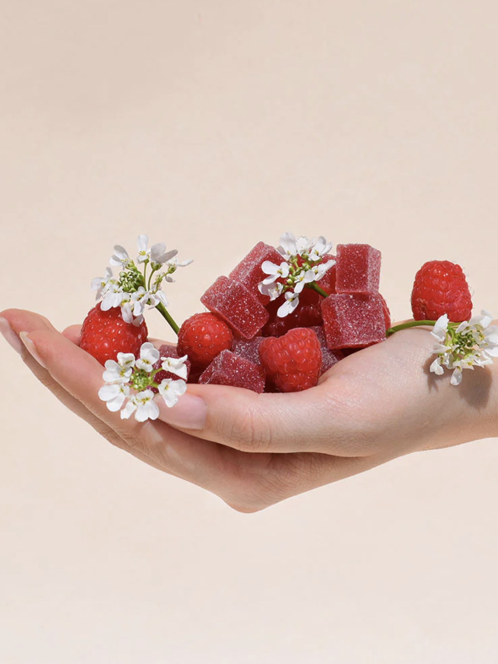 A hand holds red gummy cubes, fresh raspberries, and small white flowers against a plain, light beige background.