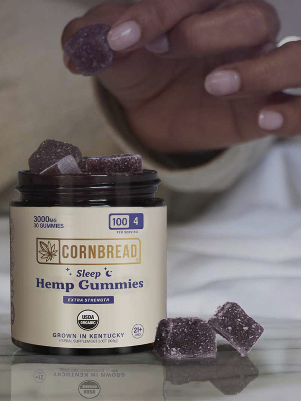 A hand picks up a purple gummy from an open jar labeled "Cornbread Sleep Hemp Gummies" on a white surface.