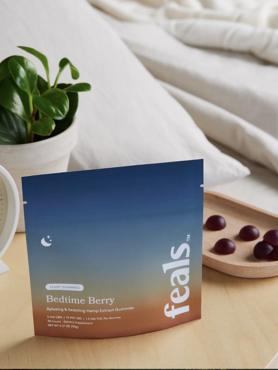 A packet of Feals Bedtime Berry sleep gummies sits on a bedside table next to a clock, a plant, and a tray with several gummies, with a bed in the background.