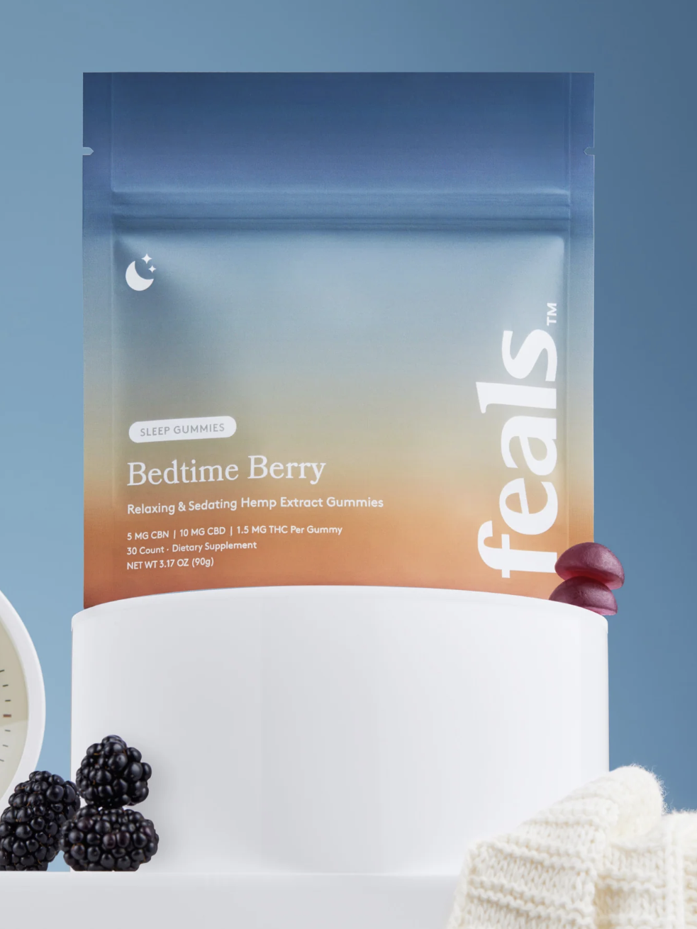 A package of Feals Bedtime Berry hemp extract gummies stands on a white pedestal next to an analog clock, blackberries, and a knit blanket against a blue background.
