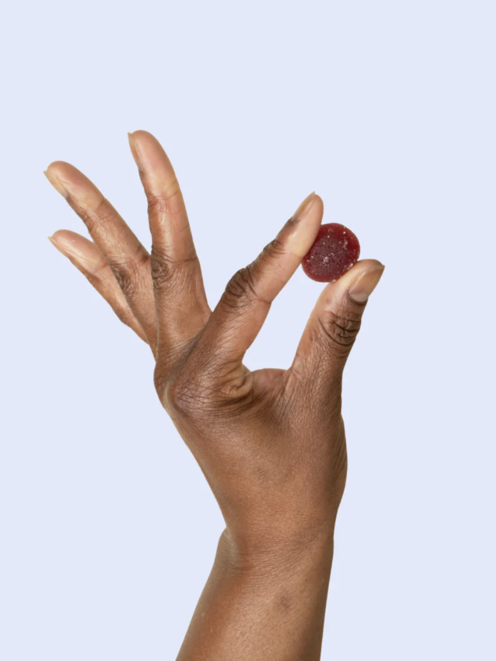 A hand with medium brown skin holds a single round red gummy candy between the thumb and middle finger against a plain light gray background.