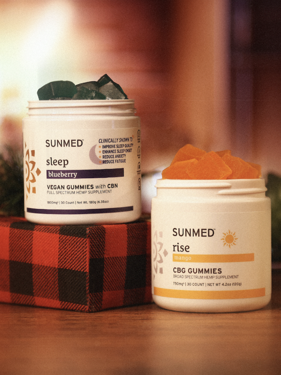 Two jars of SUNMED gummies are displayed; one labeled "sleep" with blueberry gummies and the other "rise" with mango CBG gummies, set on a red plaid surface with greenery in the background.