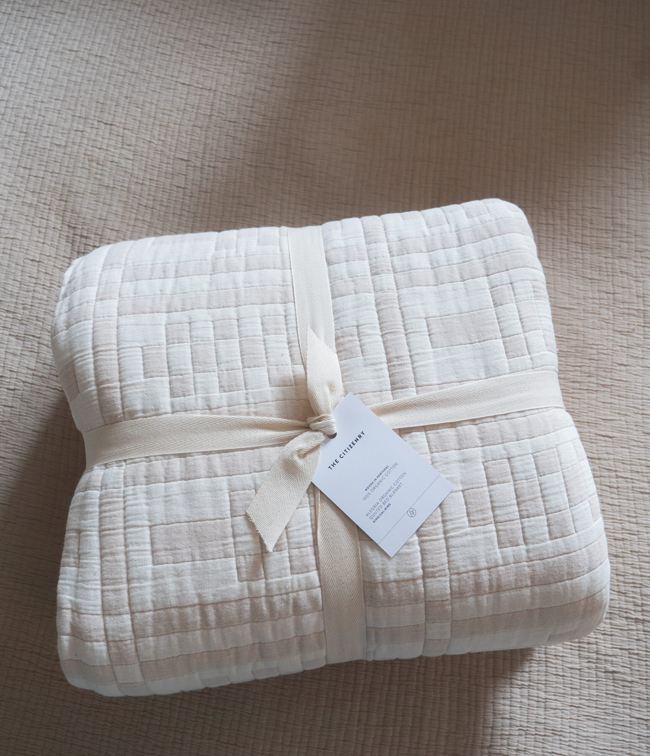 A folded cream-colored quilt with a stitched pattern, tied with a beige ribbon and a tag, placed on a textured light beige surface.
