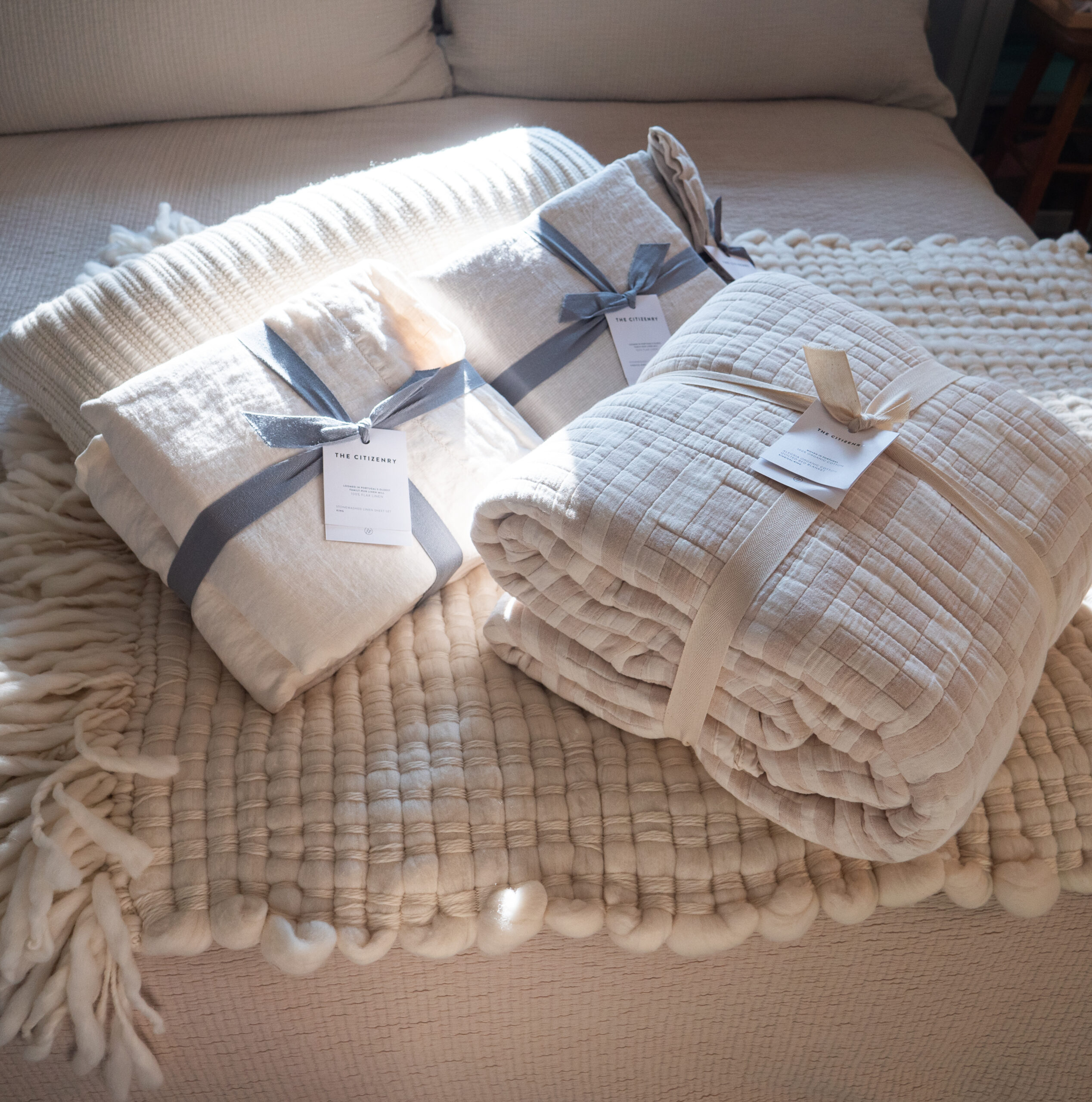 Three folded cream-colored blankets with tags and ribbon ties are placed on a textured cream bedspread in natural sunlight.