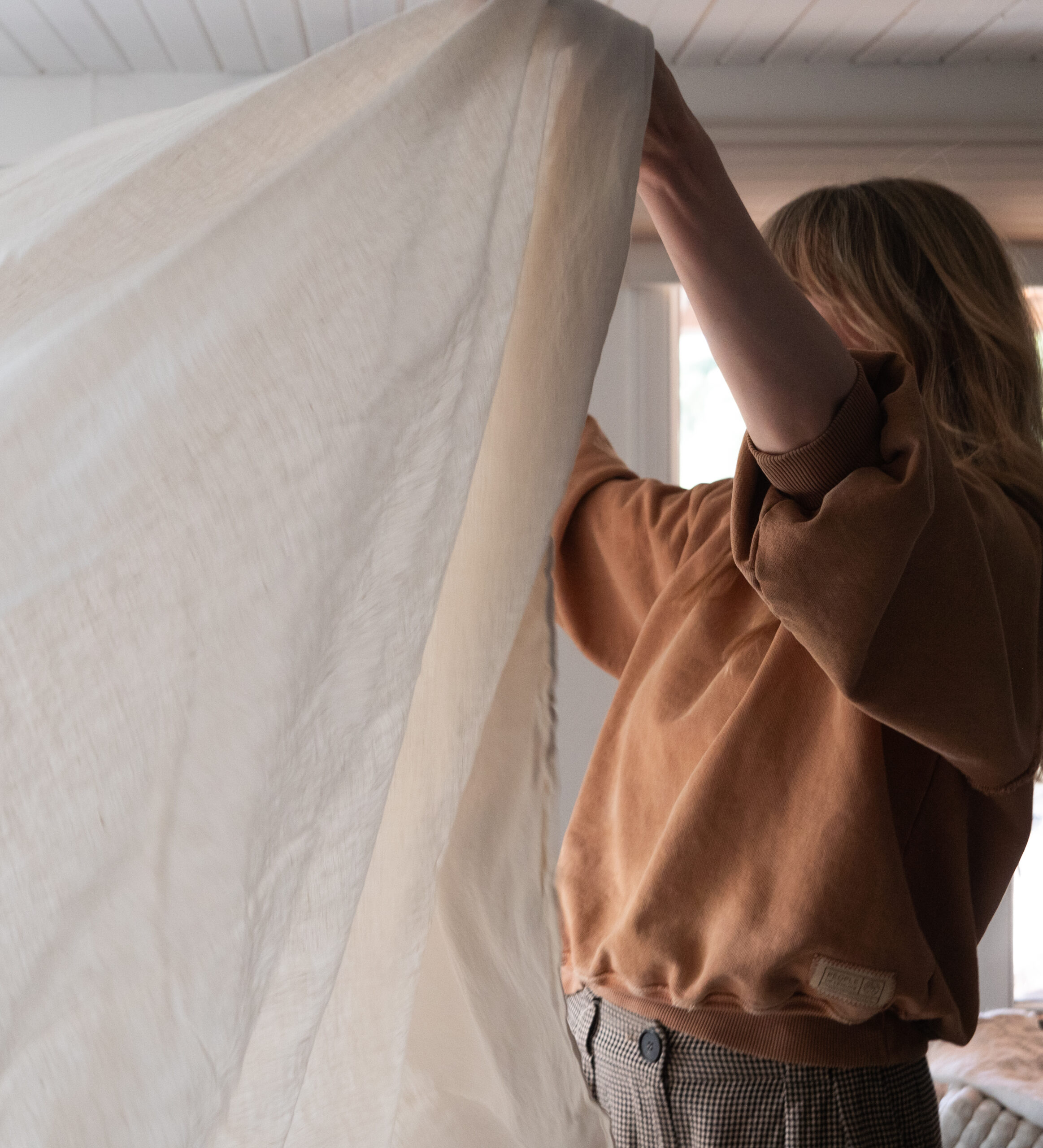 A person wearing a brown sweater holds up a large white sheet or fabric in a sunlit room.